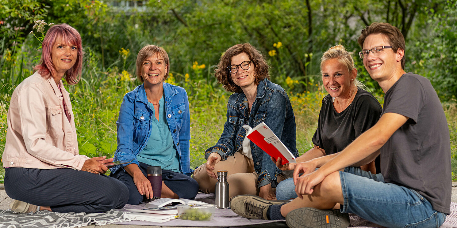 Studierende sitzen auf Decken mit ihren Lernmaterialien vor Bäumen und unterhalten sich.