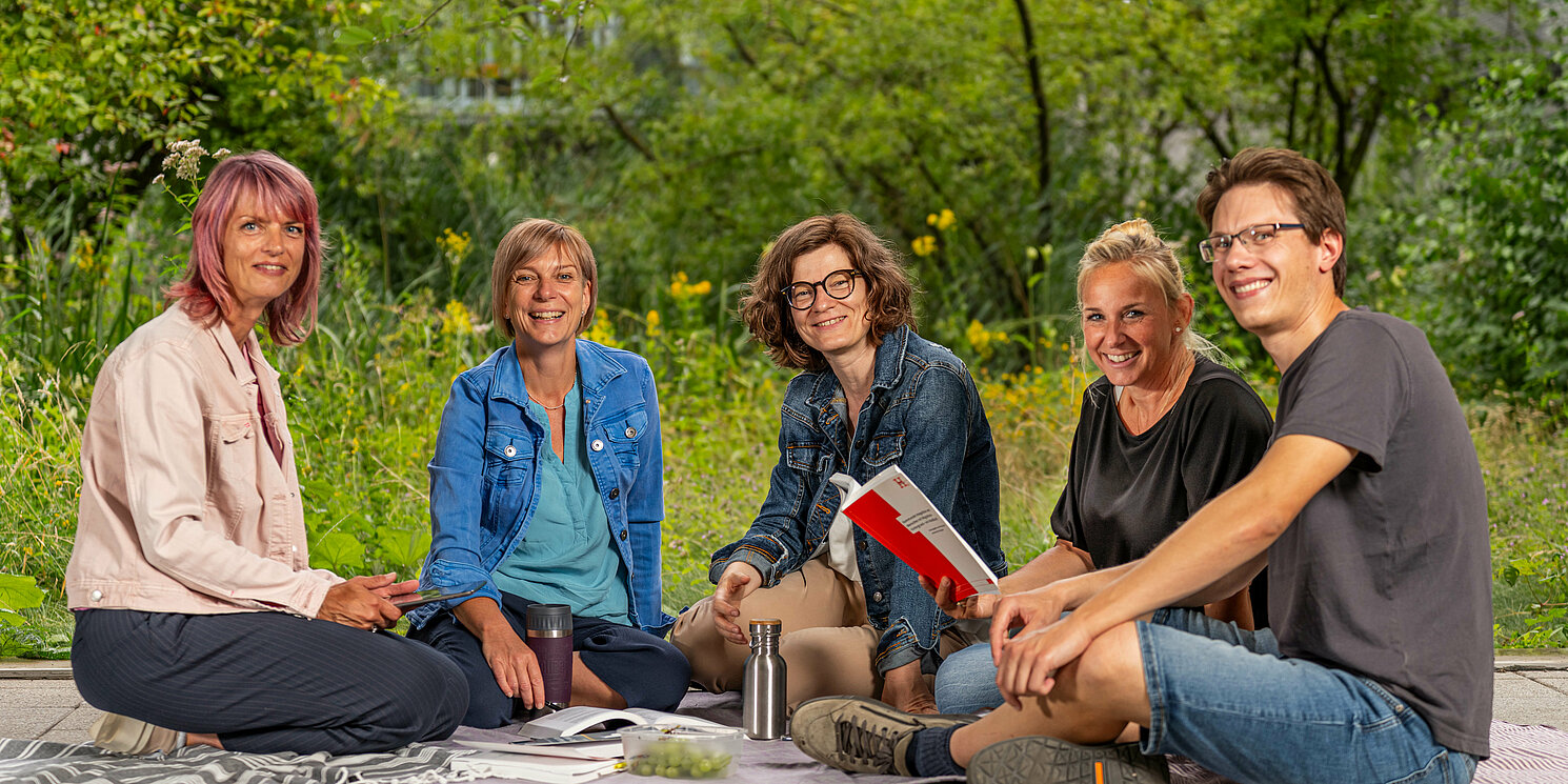 Studierende sitzen auf Decken mit ihren Lernmaterialien vor Bäumen und unterhalten sich.