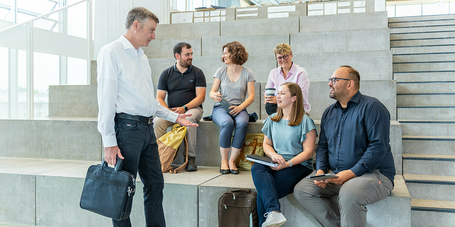Leitung und Kommunikationsmanagement berufsbegleitend studieren Studierende sitzen auf einer Treppe und unterhalten sich mit ihrem Dozenten.