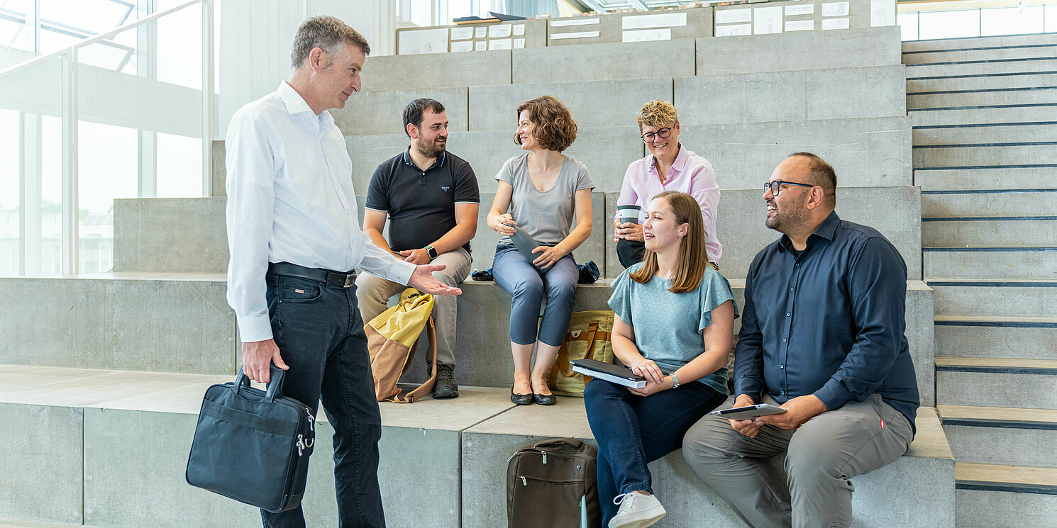 Studierende sitzen auf einer Treppe und unterhalten sich mit ihrem Dozenten.