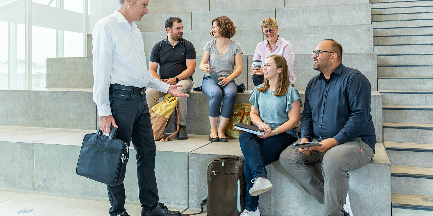 Studierende sitzen auf einer Treppe und unterhalten sich mit ihrem Dozenten.