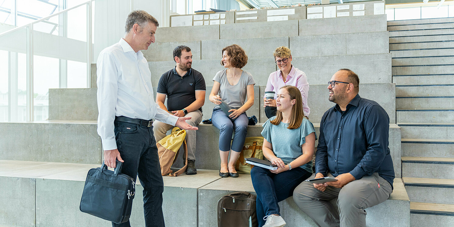 Studierende sitzen auf einer Treppe und unterhalten sich mit ihrem Dozenten.
