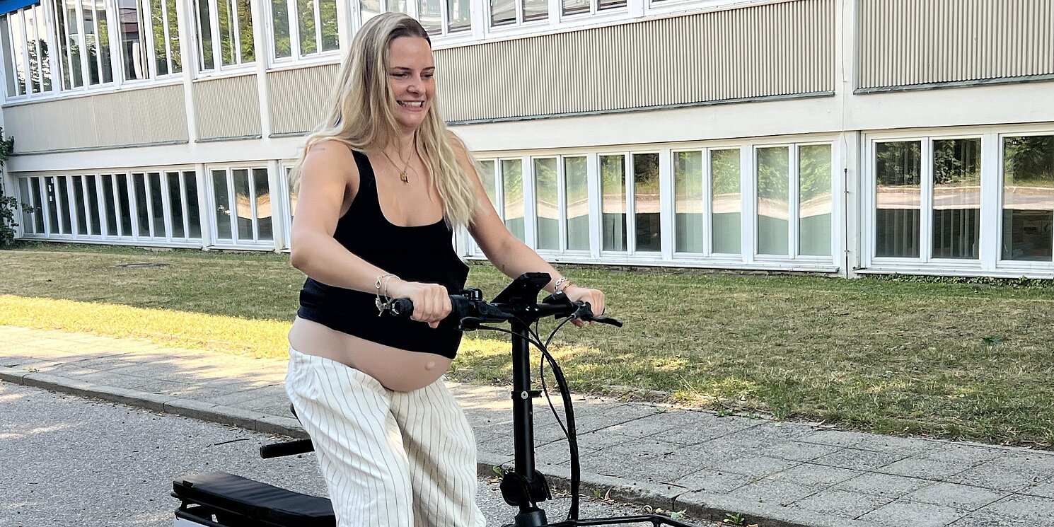 Ein Lastenfahrrad auf dem Geländer der OTH Regensburg, das von einer Studentin gefahren wird, die sich einen Schwangerschaftsbauch umgeschnallt hat. 